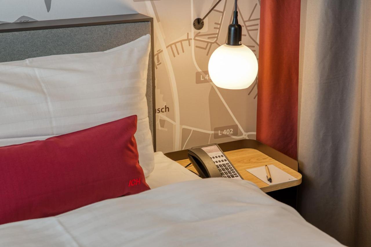 Intercityhotel Berlin Airport Ber Terminal 1 & 2 Шёнефельд Экстерьер фото The photo shows a cozy bedroom setup. There's a neatly made bed with white bedding and a red decorative pillow. Beside the bed, a small bedside table holds a telephone and a notebook or notepad, along with a pen. A round pendant light hangs above the